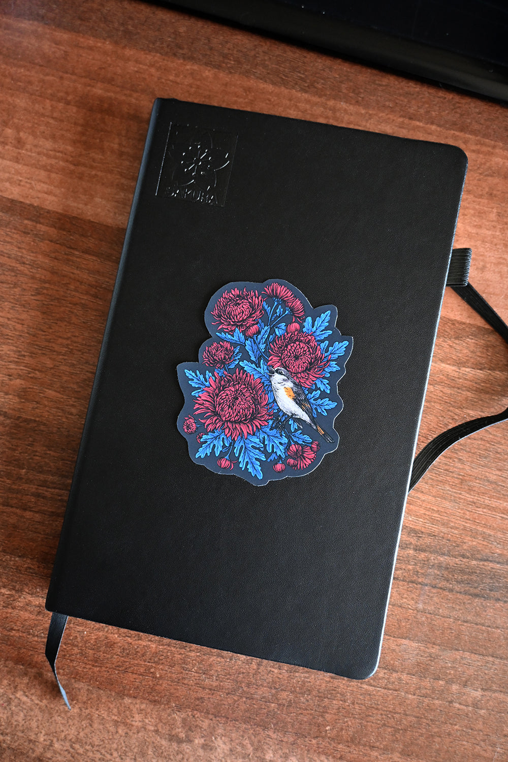 Black notebook with a vinyl sticker with red mums and a restart bird on a wooden surface