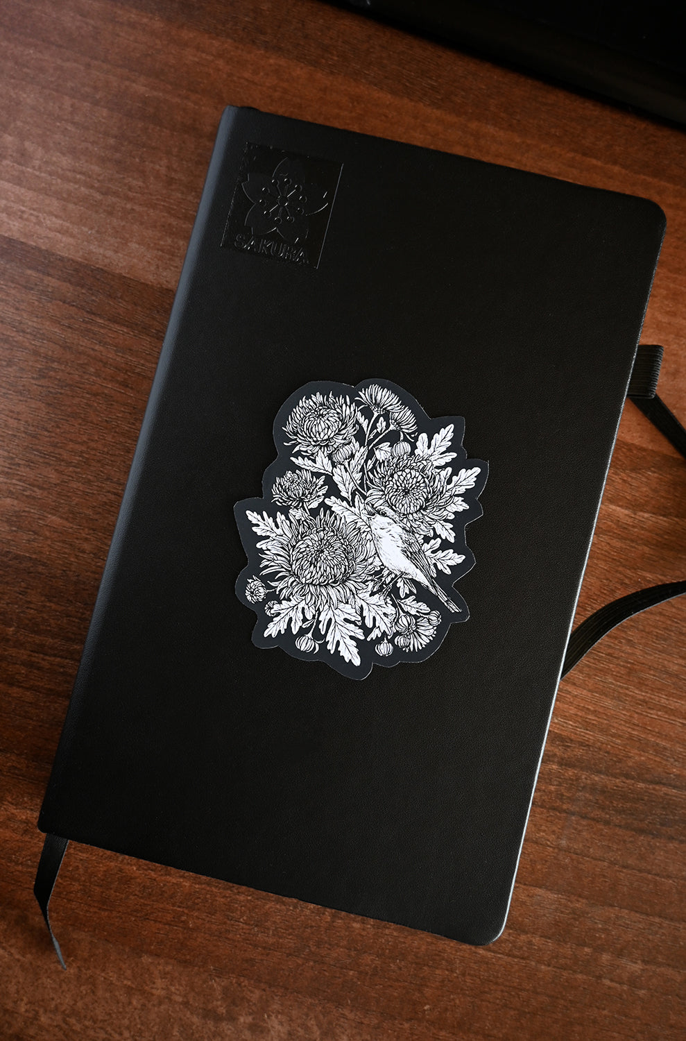 Mums and redstart bird, black and white vinyl sticker on a black notebook laying on a wooden surface