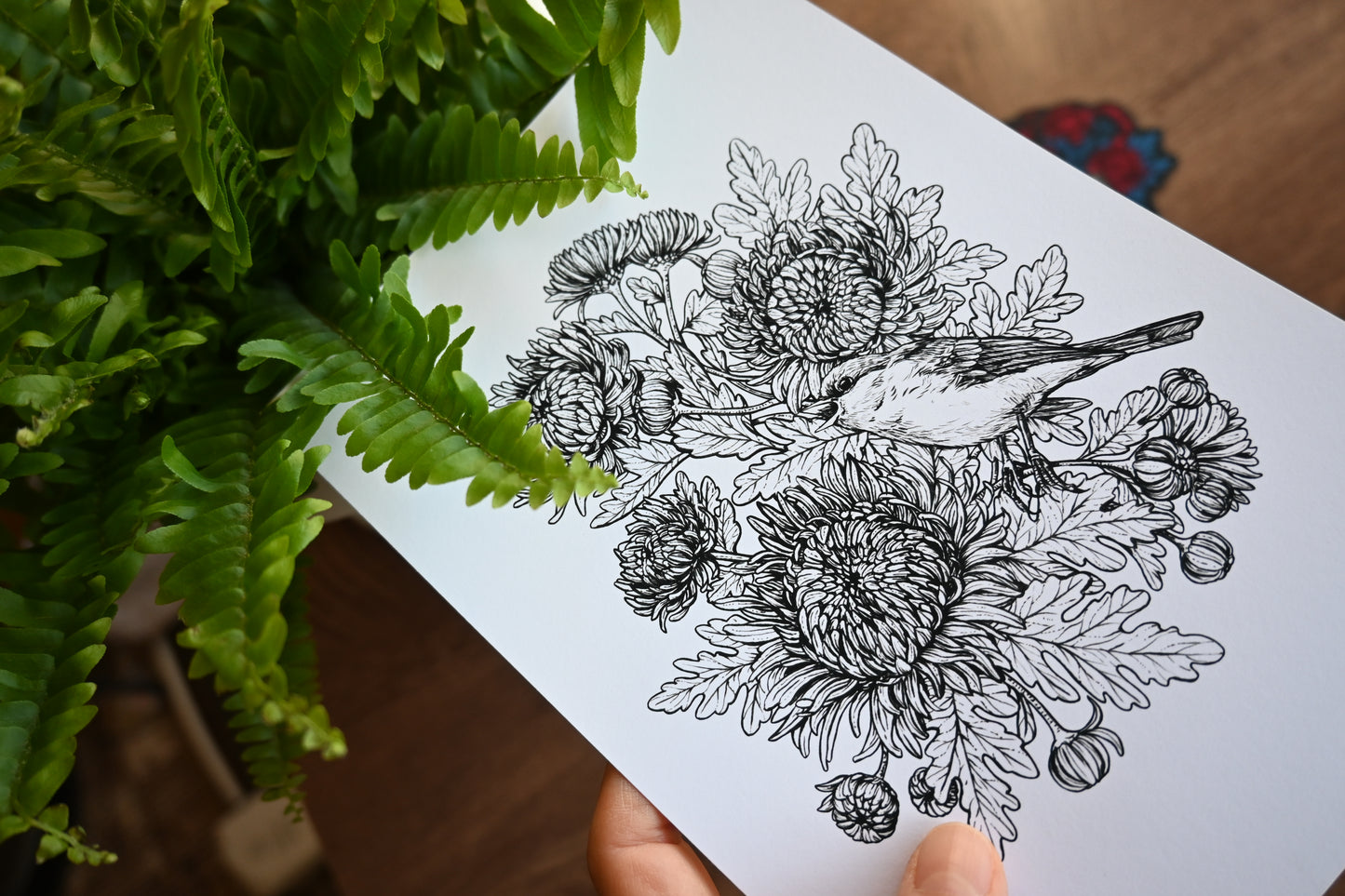 Hand holding a card with black floral and bird illustration against a green leafy background