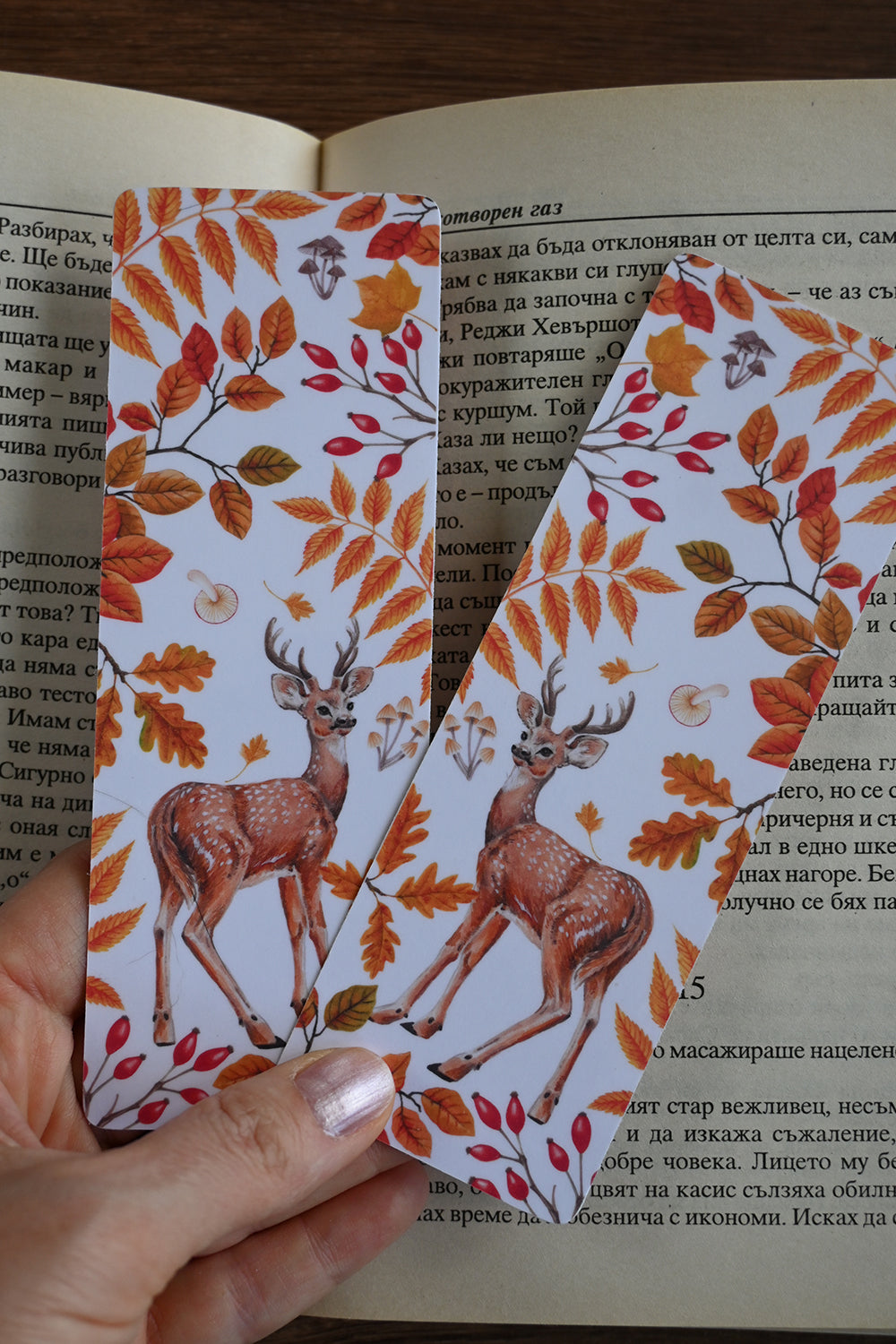 Two deer-themed bookmarks with autumn leaves held over an open book.