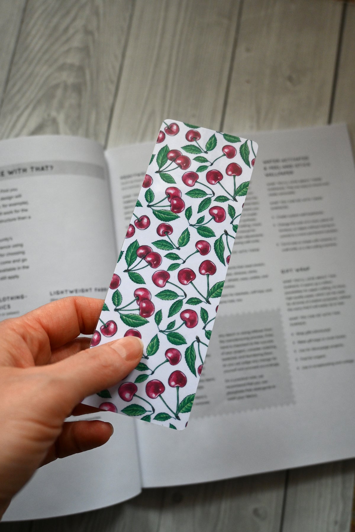 Hand holding a cherry-patterned bookmark over an open book on a wooden surface