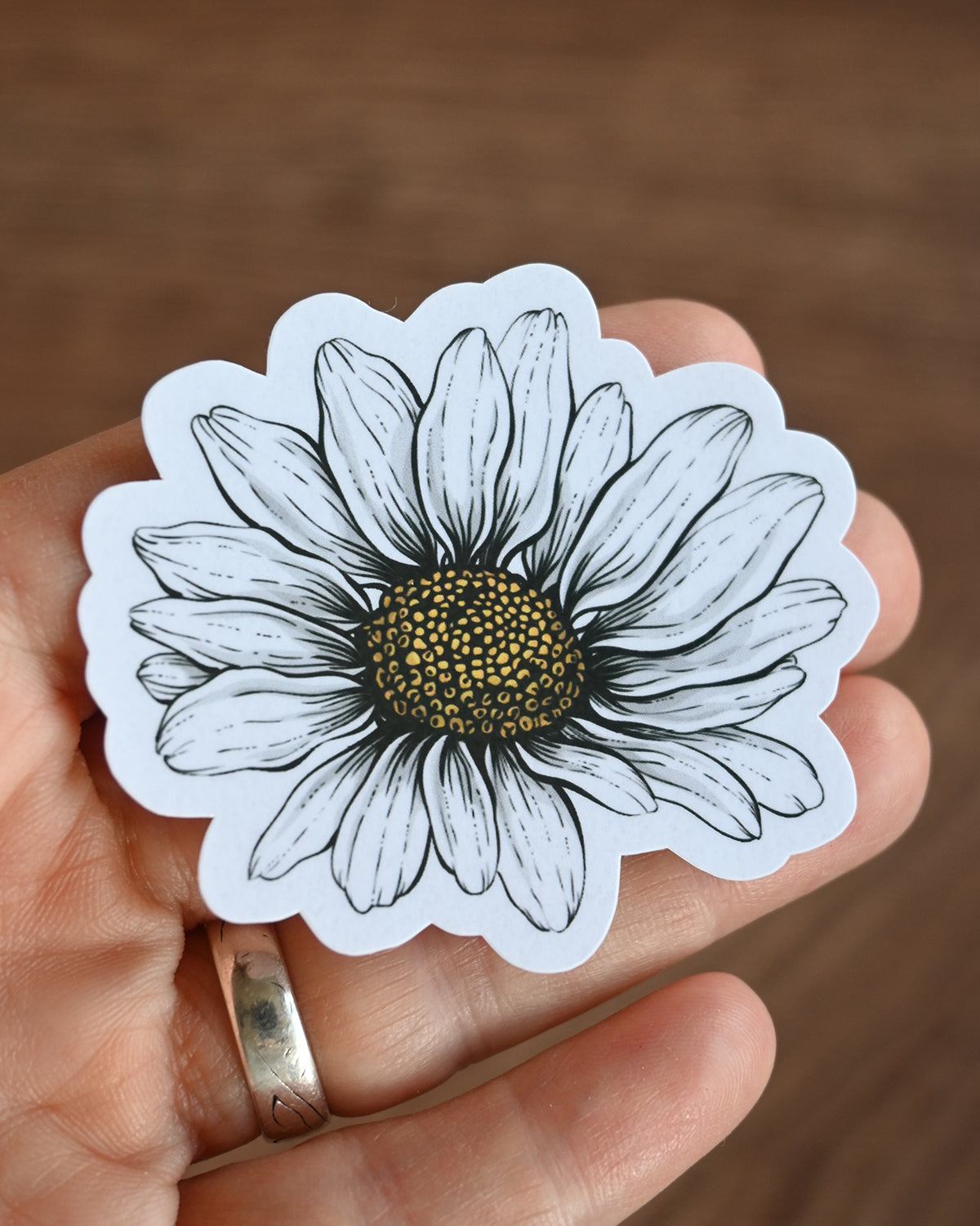 Hand holding a white sticker with a daisy design on a brown background