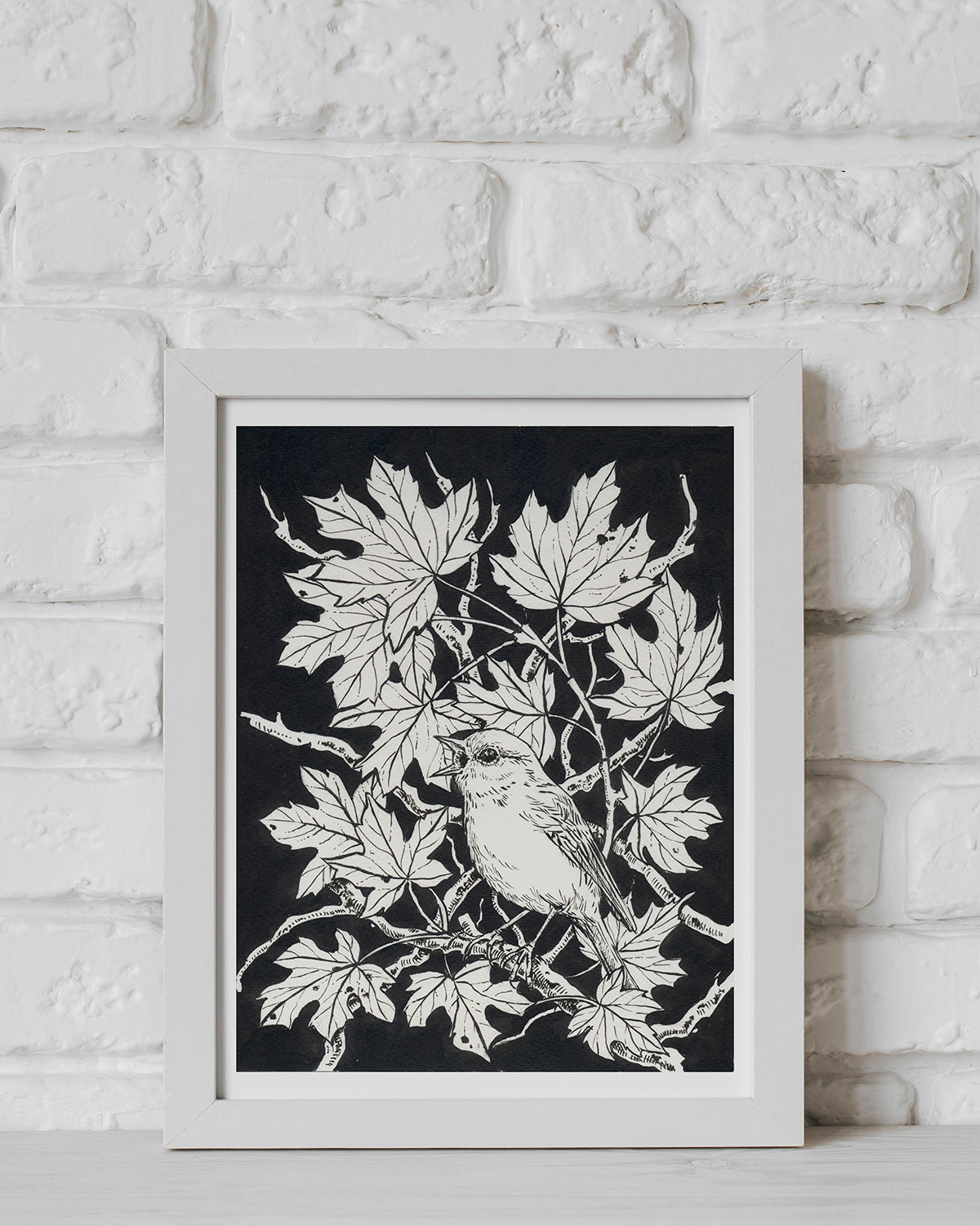 Framed artwork of a bird on a branch with leaves against a black background, displayed on a white brick wall.