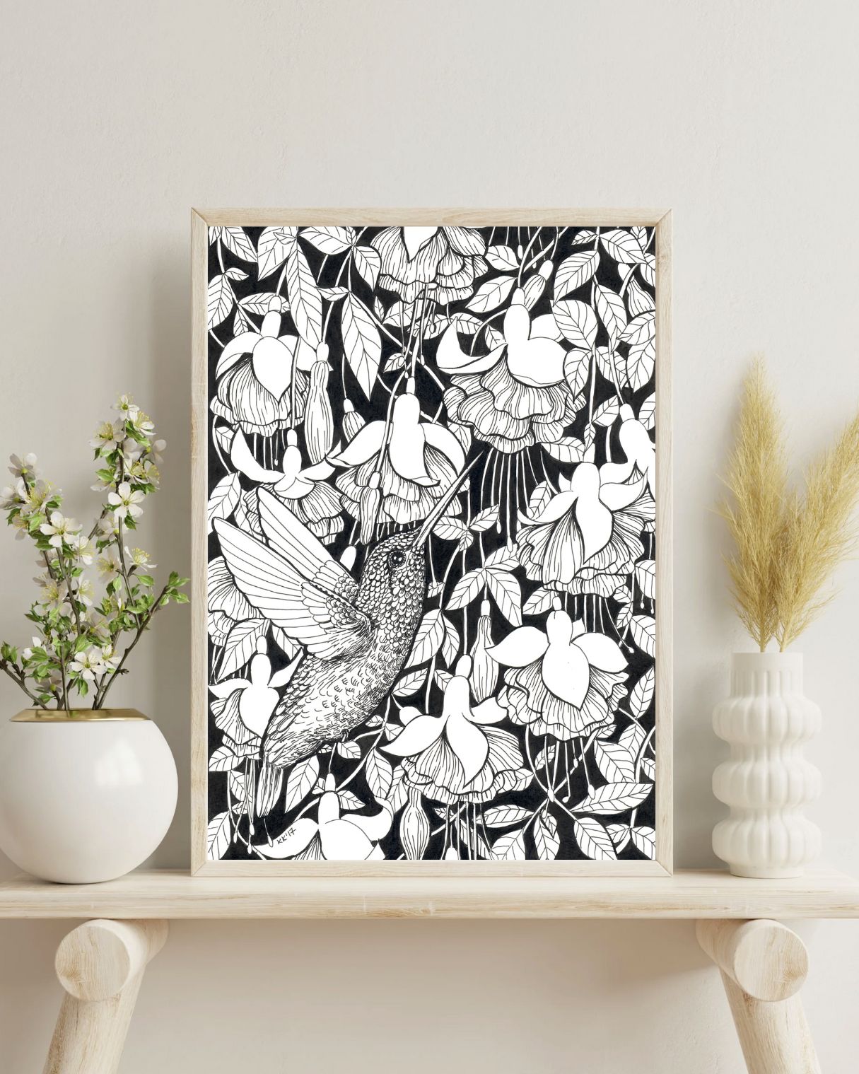 Framed black and white ink drawing of a hummingbird and fuchsia on a shelf with decorative items.