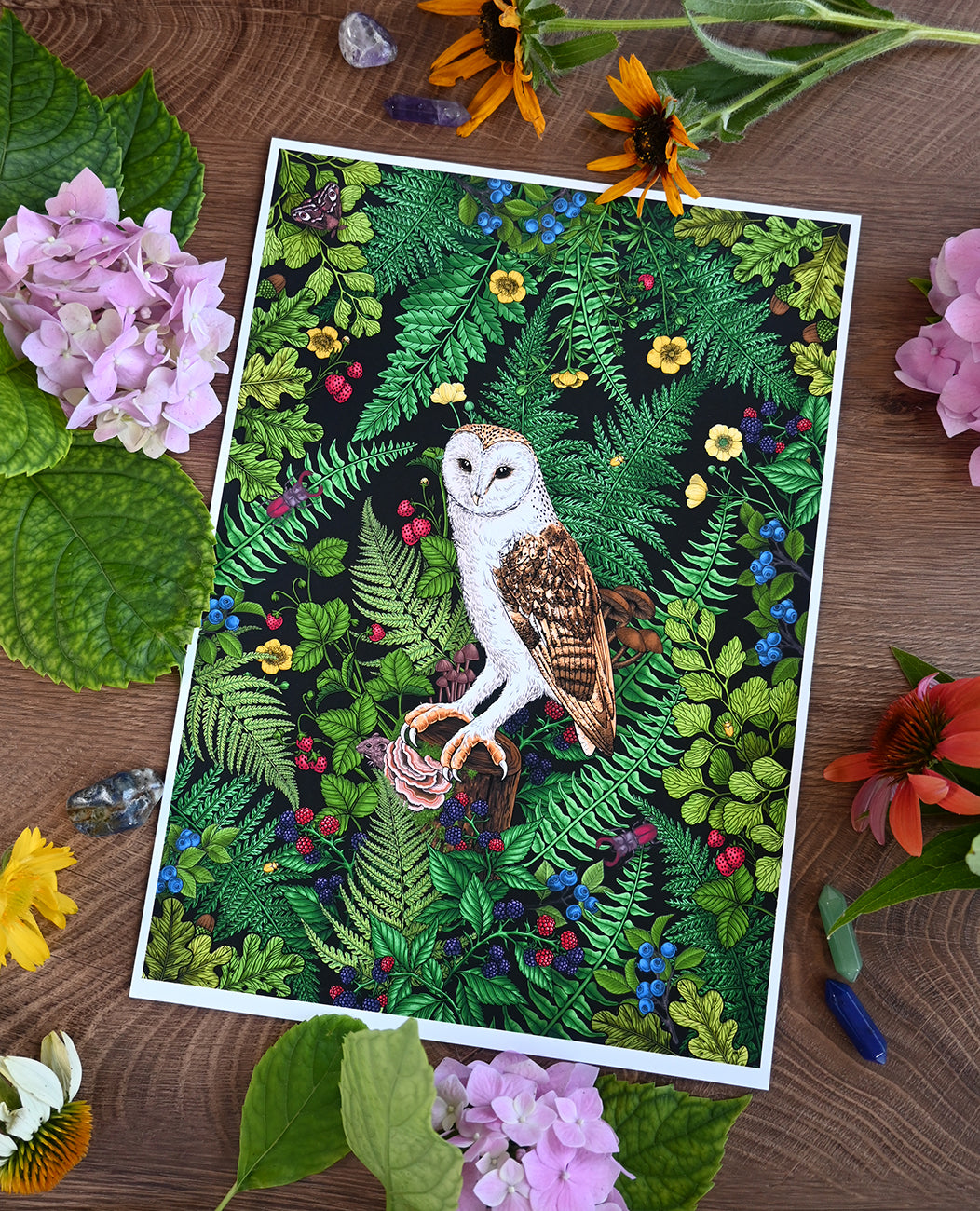 Print of an illustration of an owl in a forest setting surrounded by flowers and leaves on a wooden surface.