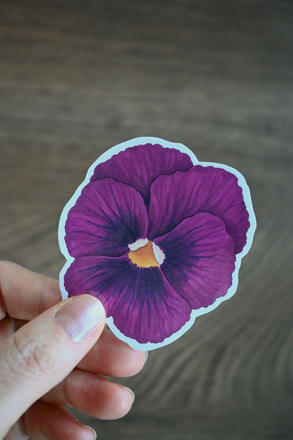 Hand holding a purple pansy sticker against a neutral background