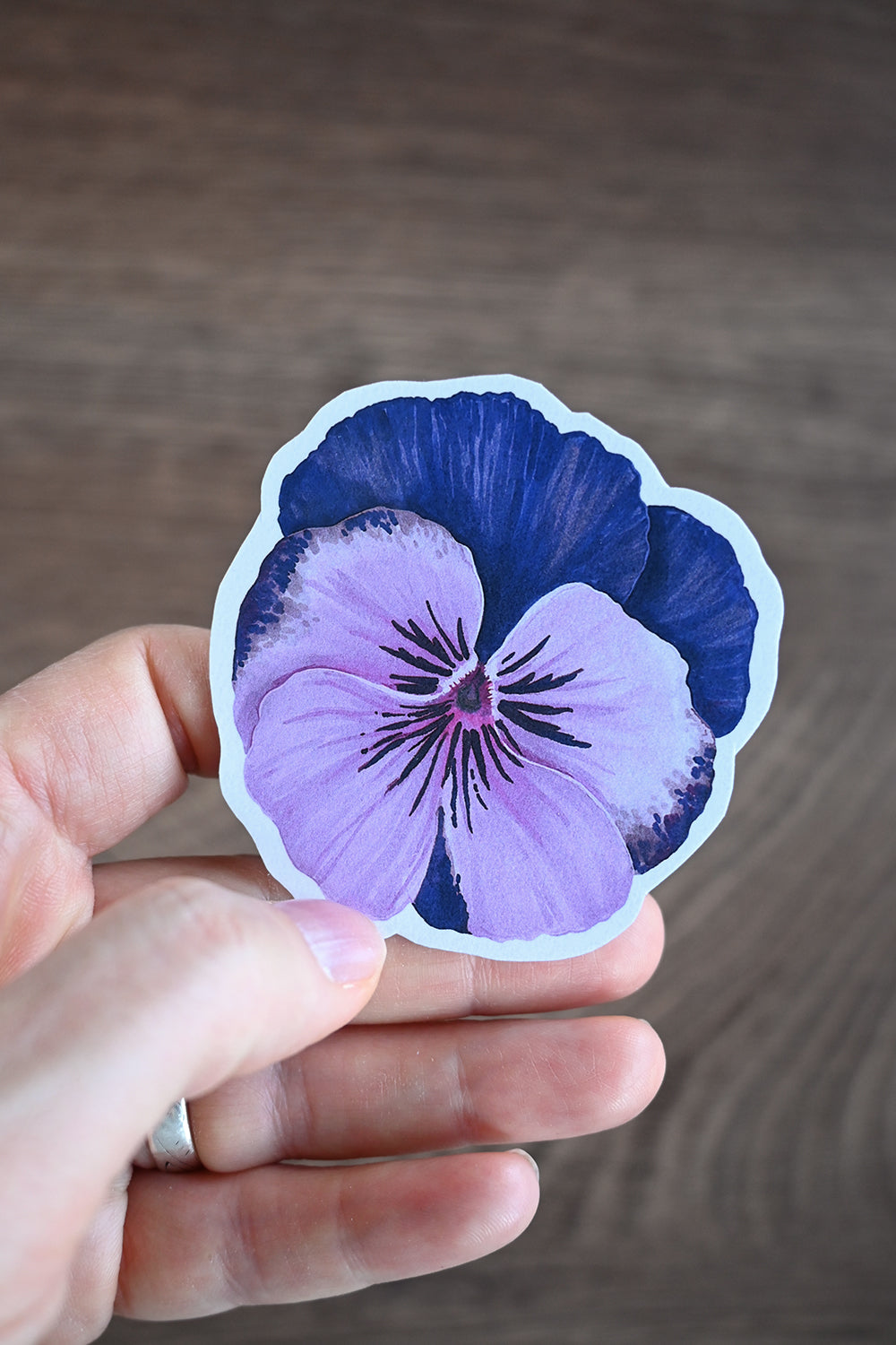 Hand holding a purple and blue pansy sticker against a wooden background