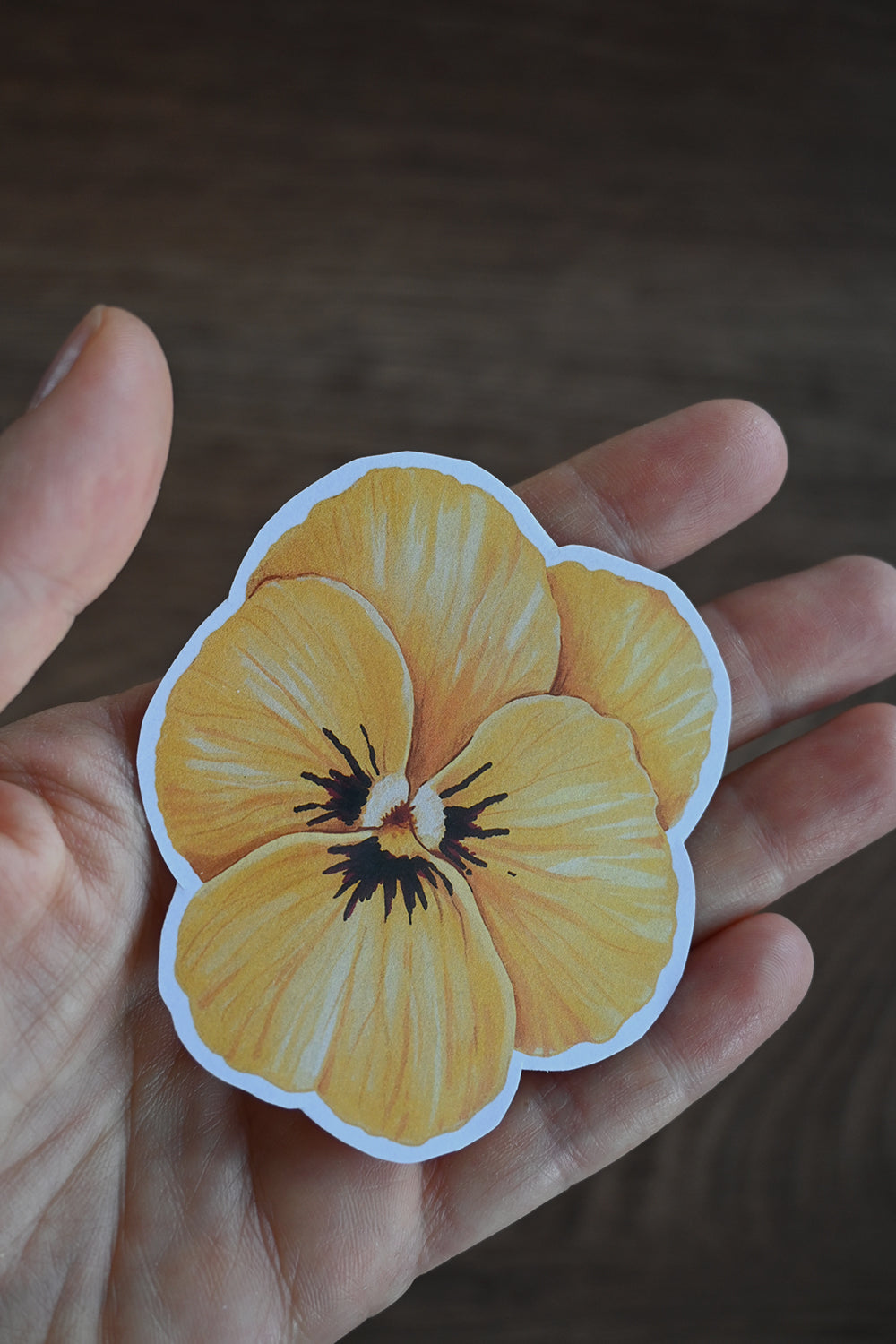 Hand holding a yellow pansy sticker against a blurred background