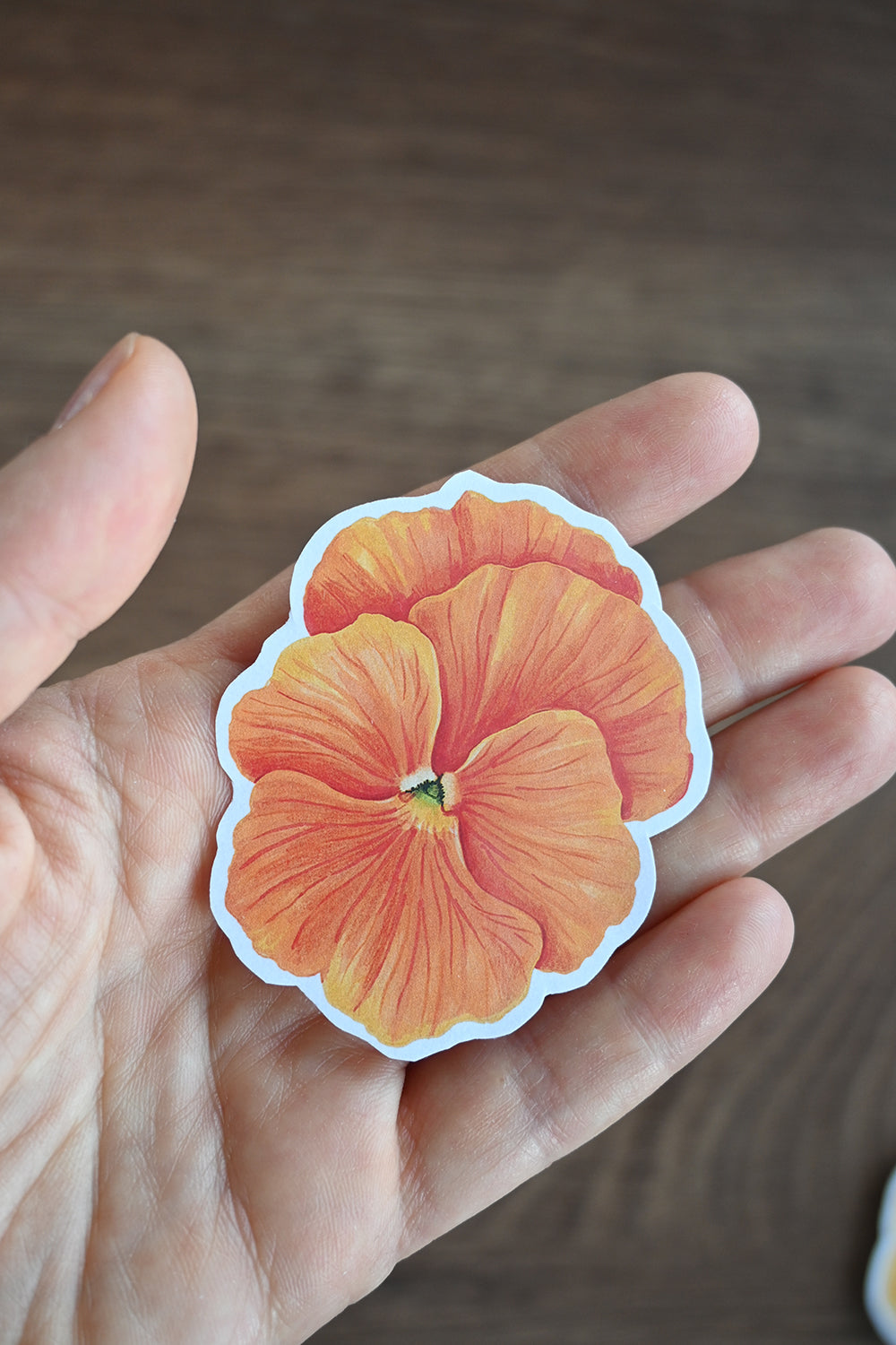 Hand holding an orange pansy sticker against a blurred background