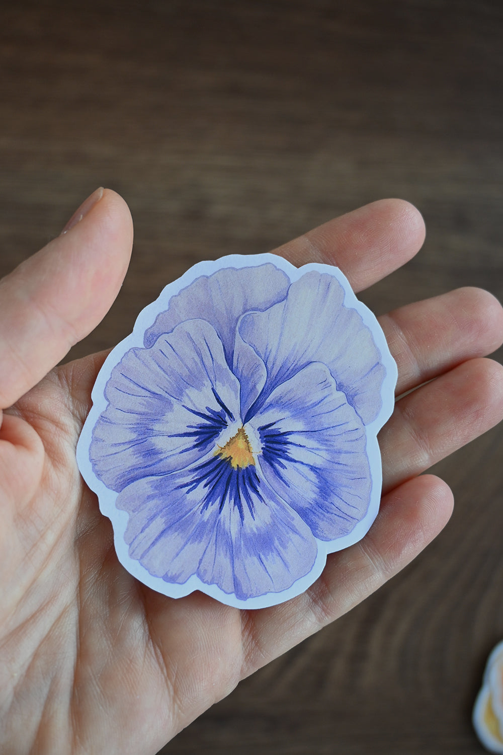 Hand holding a light blue pansy sticker against a blurred background