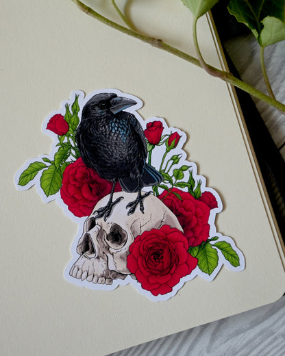 photo of a vinyl sticker with printed illustration of a raven perching on a human skull with red roses behind