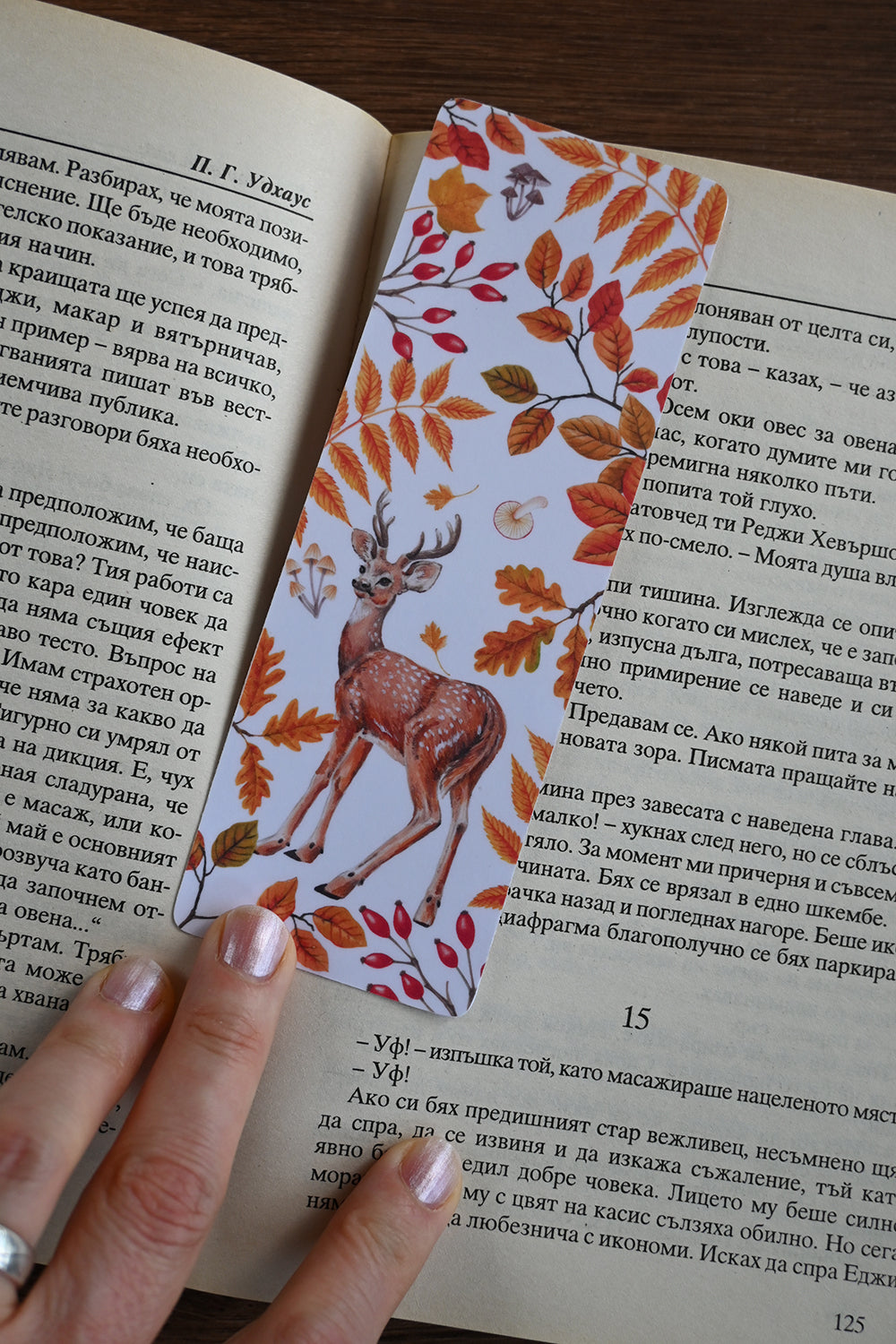 Open book with a decorative bookmark featuring a deer and autumn leaves, held by a hand.