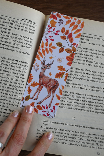 Open book with a decorative bookmark featuring a deer and autumn leaves, held by a hand.