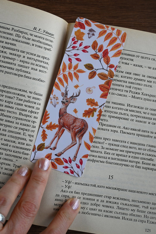 Open book with a decorative bookmark featuring a deer and autumn leaves, held by a hand.