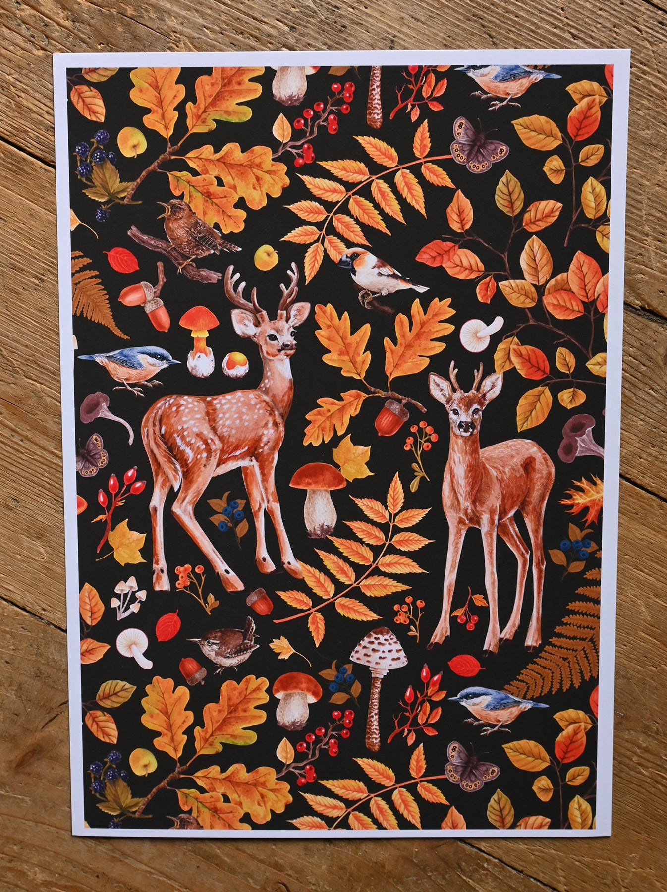 print of two deer among autumnal flora, mushrooms and birds on wooden surface