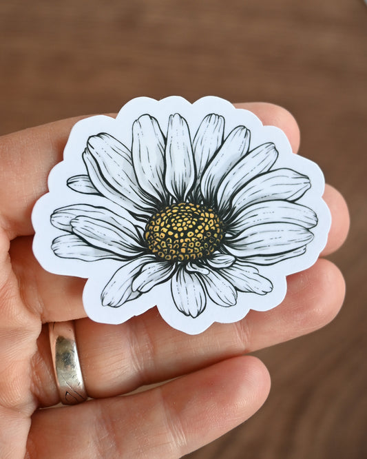 Hand holding a white sticker with a black and white flower design against a brown background