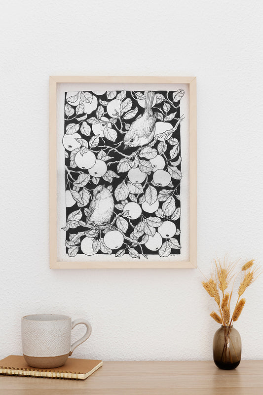 Framed black and white artwork of birds and fruit on a wall above a desk with a mug and vase.