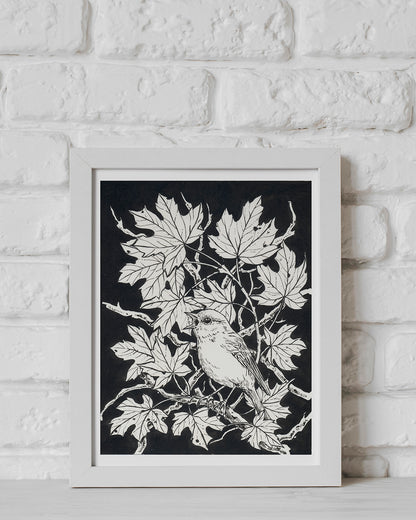 Framed artwork of a bird on a branch with leaves against a black background, displayed on a white brick wall.