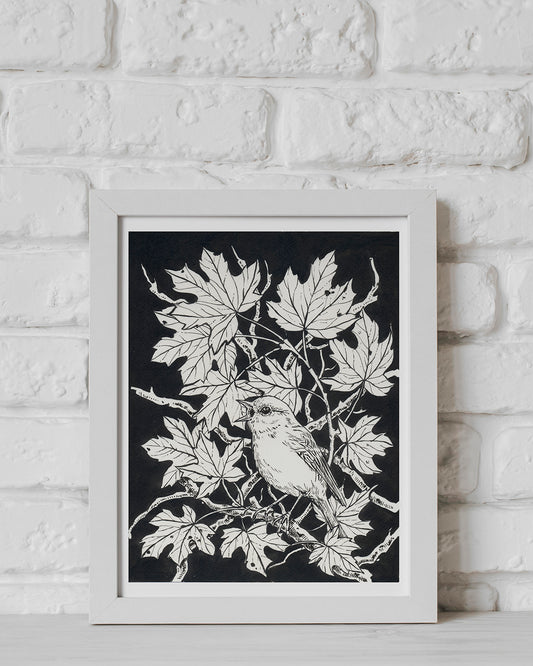 Framed artwork of a bird on a branch with leaves against a black background, displayed on a white brick wall.