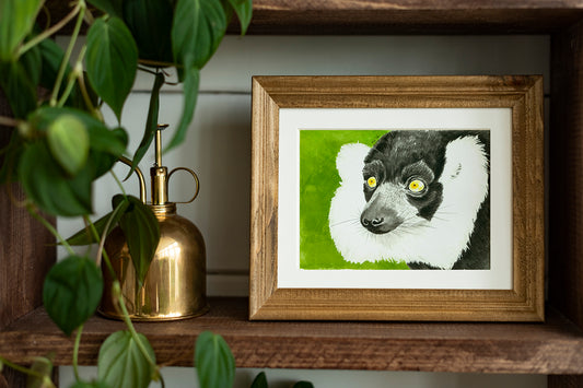 Framed artwork of a lemur on a wooden shelf with plants and a gold lamp.