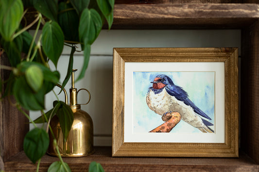 Framed artwork of a swallow on a wooden shelf with a plant and a brass container.